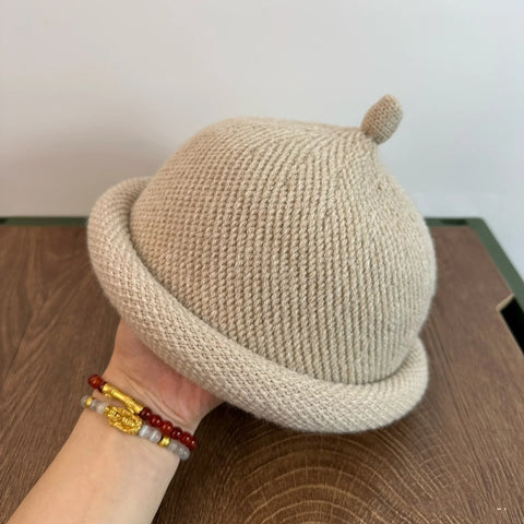 Beige knit hat with a small pom-pom held by a hand on a wooden surface.